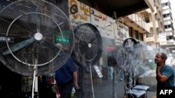 资料照片：2021年6月30日，在伊拉克首都巴格达，一名男子站在风扇旁边，风扇上喷洒着捐助者提供的混合水蒸气，为街道上的行人降温。