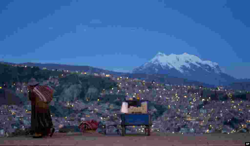 A woman walks along Pereferica Avenue, past a street stall of popcorn, known as "pipocas," backdropped by the snow-capped Illimani Mountain, at sunset in La Paz, Bolivia.
