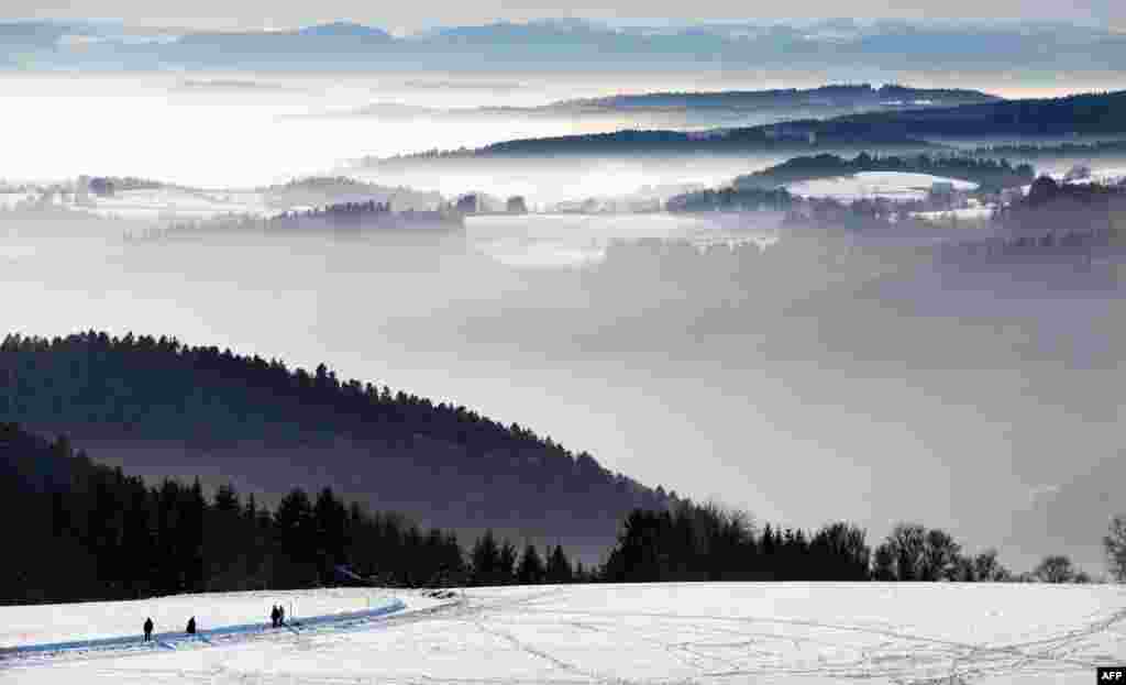 People walk on a snowy landscape as the Swiss Alps are seen in the background and mist partly covers the alpine upland near the southern German town of Höchenschwand.