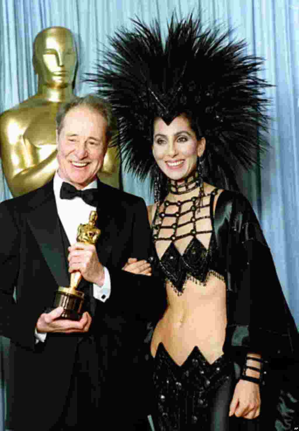 Nữ ca sĩ kiêm diễn viên Cher xuất hiện tại lễ trao giải Oscar năm 1986 với phong cách thời trang gây sốc. Bên cạnh là nam diễn viên Don Ameche với tượng vàng cho Nam diễn viên phụ xuất sắc nhất.” (AP)