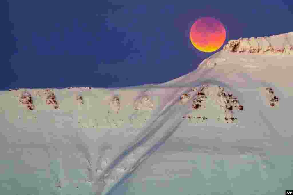 2018年1月31日，从挪威斯瓦尔巴群岛的朗伊尔城（Longyearbyen）可以看到雪山之巅的红黄相间的明月。